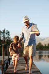 Grandfather grandson with toy sailboat walking along dock over lake - obrazy, fototapety, plakaty