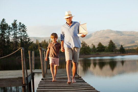 Smiling Grandfather Grandson Toy Sailboat Holding Hands Walking Along Dock Over Lake