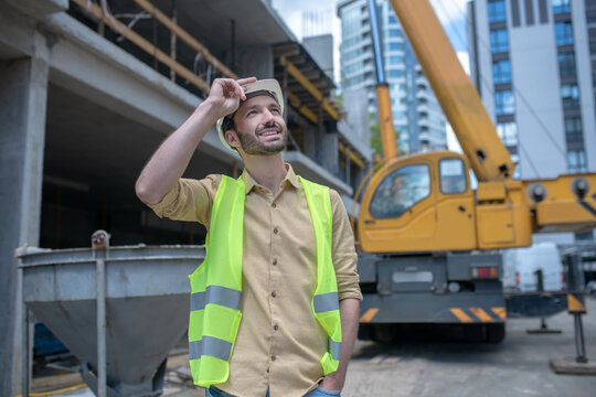 Building Worker In Helmet And Yellow Vest Touching His Helmet With His Hand, Looking Up