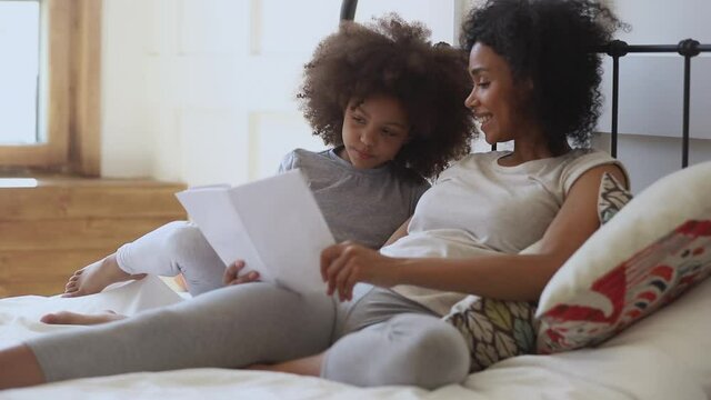 Cute little african kid daughter learning to read telling fairy tale to happy black mother holding book relaxing in bed, funny small child girl enjoying bedtime stories talking to mom in bedroom