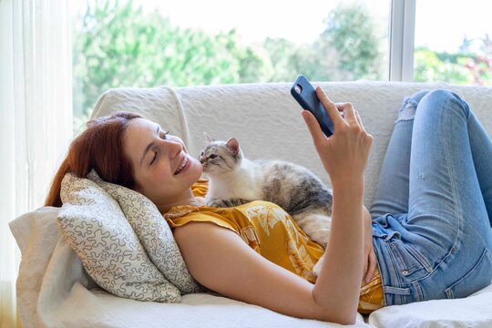Happy young woman lying on the couch at home with her cat using smartphone