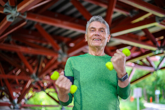 Low angle portrait of smiling senior man exercising with dumbbells while standing at gazebo - Powered by Adobe
