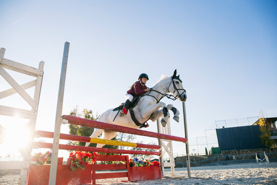 Low angle view of teenage jockey girl riding white horse over hurdle on training ground against clear sky - Powered by Adobe
