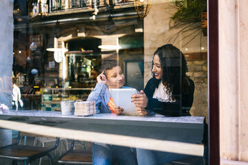 Smiling boy looking at mother sitting with digital tablet in restaurant seen through window