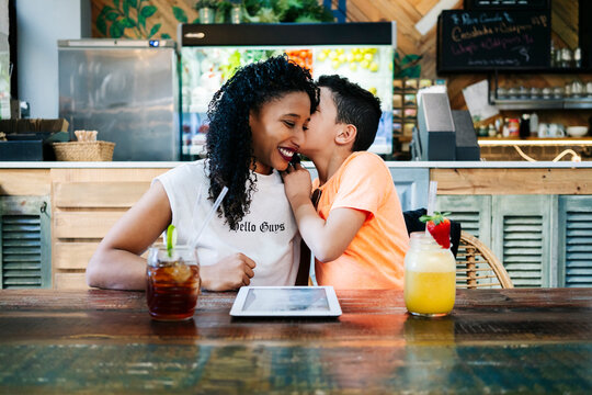 Boy whispering in smiling mother's ear while sitting with drinks at restaurant