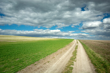 A dirt road through rural fields on the hill and grey clouds on the blue sky