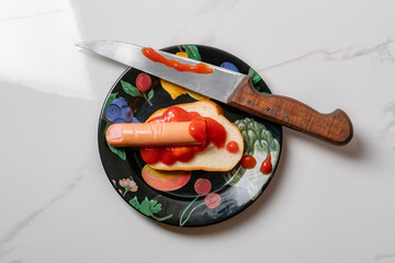 Sausage in ketchup on a piece of bread next to a knife. Sausage in the shape of a bloodied finger.