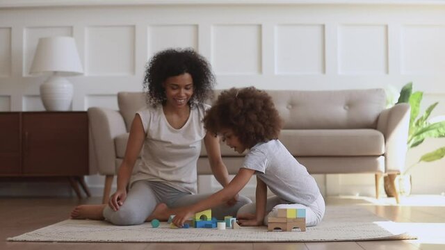 Happy black family of two cute little african american kid daughter and mother playing on floor, smiling mom helping small preschool child girl building constructor castle of wooden blocks at home