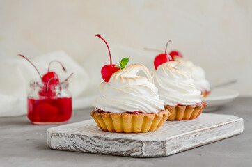 Tartlets with cherry filling and Italian meringue with a cocktail cherry on top on a board on a concrete background. Horizontal orientation. Food background.