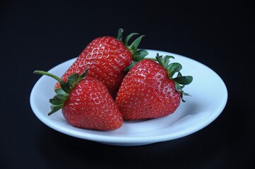 Three ripe red juicy strawberries on a white saucer on a black background