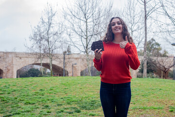 Portrait of pretty caucasian young woman with chocolate  showing thumb up gesture, in the park, orange sweater and jeans. Place for your text in copy space.