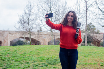 happy attractive caucasian young woman holding rotating and styling brush, taking a selfie, in the park, orange sweater and jeans, long curly hair. Place for your text in copy space.