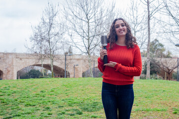 Portrait of attractive caucasian young woman model holding rotating and styling brush, in the park, orange sweater and jeans. Place for your text in copy space.