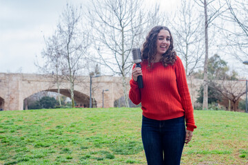 Portrait of happy attractive caucasian young woman holding rotating and styling brush  winking, in the park, orange sweater and jeans, long curly hair. Place for your text in copy space.