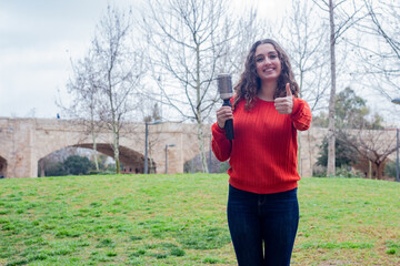 Portrait of pretty young woman model with rotating and styling brush  showing thumb up gesture, in the park, orange sweater and jeans, long curly hair. Place for your text in copy space.