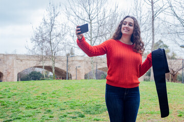 happy attractive caucasian young woman model holding electronic abdominal appliance, taking a selfie, in the park, orange sweater and jeans, long curly hair. Place for your text in copy space.