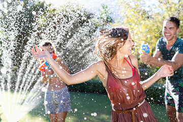 Friends playing with water guns in sprinkler - obrazy, fototapety, plakaty
