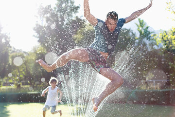 Father and son playing in sprinkler in backyard - obrazy, fototapety, plakaty