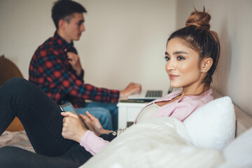 Fototapeta premium Young caucasian couple sitting in the bedroom and using modern devices working and spending time together