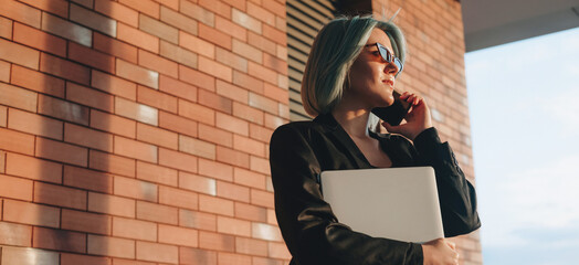 Caucasian business director talking on phone outside while holding a laptop in a brick wall