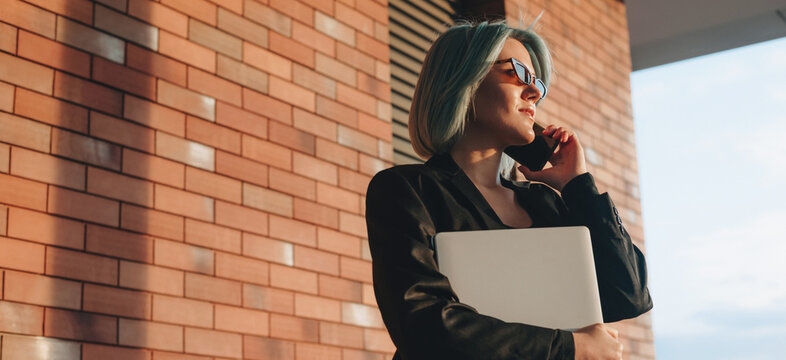 Caucasian business director talking on phone outside while holding a laptop in a brick wall