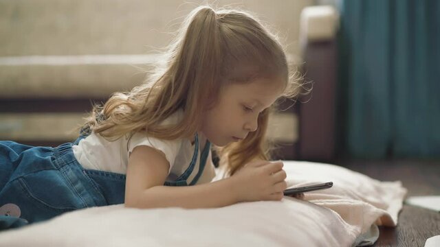 concentrated blonde little girl in denim dress plays with mobile phone lying on floor with pillow and blanket in light room at quarantine time