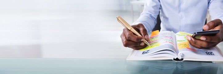 Businessman Holding Cellphone Writing Schedule In Diary With Pen
