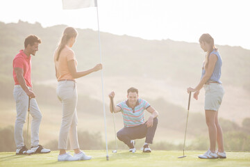 Friends watching man prepare to putt on golf course - obrazy, fototapety, plakaty
