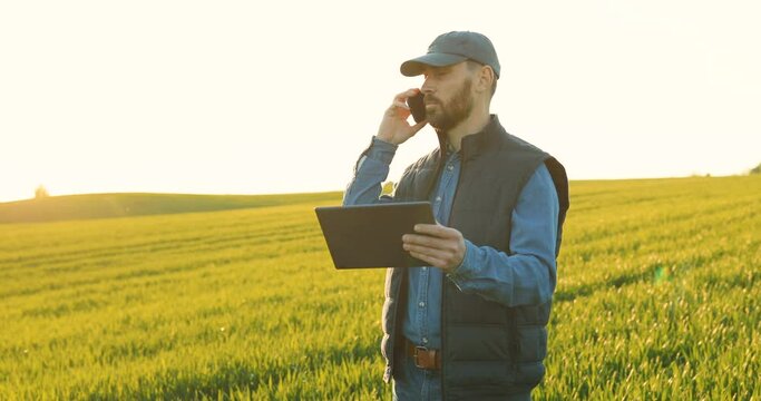 Caucasian young male farmer in hat talking on mobile phone in field. Attractive man speaking on cellphone in margin in summer and holding tablet device in hand. Looking at computer screen.