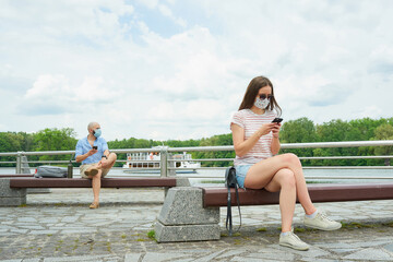 Woman on a bench keeping a social distance with a man sitting on another bench on the promenade. Girl and guy in face masks having a rest in social distancing against the background of a floating ship