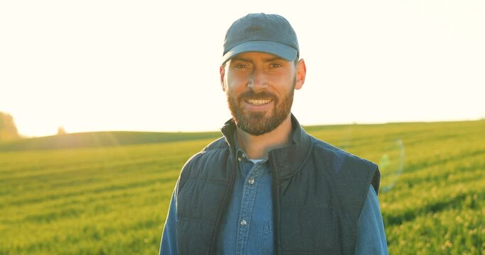 Portrait shot of handsome young Caucasian man in hat and jeans shirt standing in green field and smiling joyfully to camera. Close up of male farmer outdoor in summer. Camera zooming in on smiled face