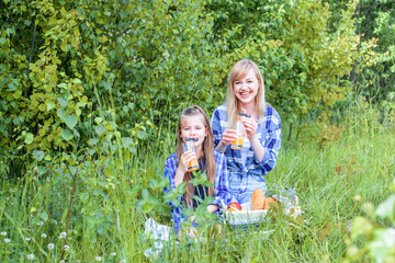 Outdoors park picnic. Beautiful young mom and little girl sitting in forest and enjoying.white bread, pastries, fruits in basket, apples and bananas.Drink orange juice from glass from straw. mustache.