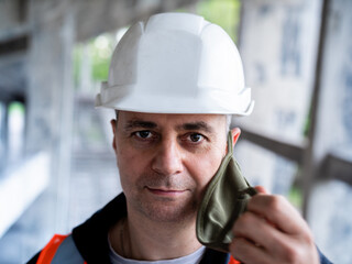 Portrait of a male Builder in a construction helmet and a medical antiviral mask. The man removes the protective mask from his face to symbolize the end of the pandemic.