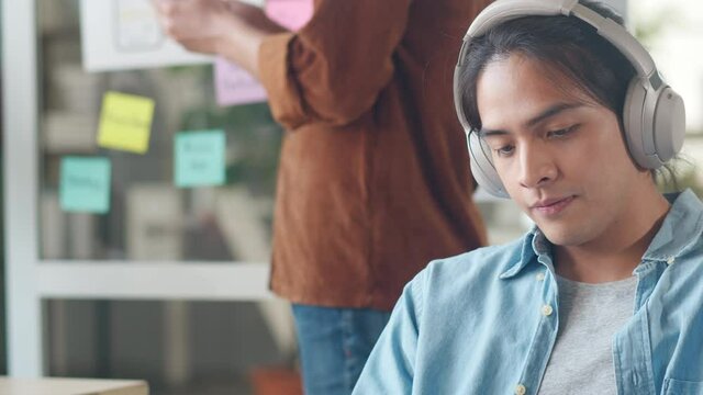 Young Asian designer man listening to music on headphones, looking at camera and smiling in modern office. Group of young college students in smart casual wear on campus. Coworker teamwork concept.
