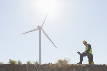 Worker using laptop by wind turbine in rural landscape