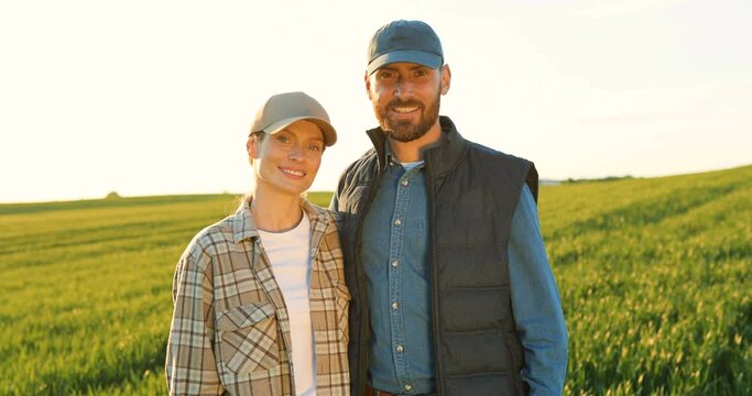 Portrait of happy couple of farmers standing in green field together and smiling cheerfully. Pretty woman and handsome man hugging in margin with wheat agriculture. Family of organic food producers.
