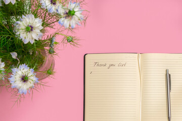 Above view of spring flowers and open gratitude journal with handwritten words Thank you list on soft pink background