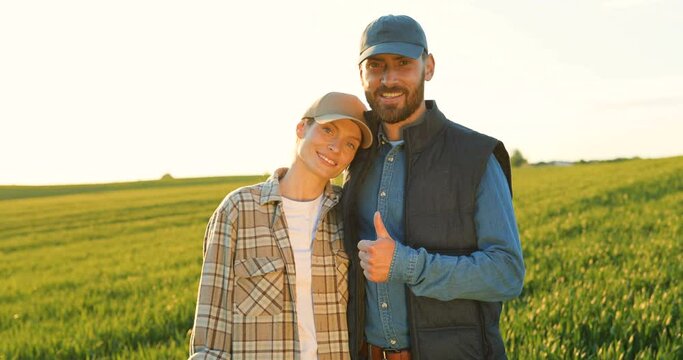 Portrait of happy couple of farmers standing in green field together and smiling cheerfully. Pretty woman and handsome man hugging in margin and giving thumb up. Family of organic food producers.