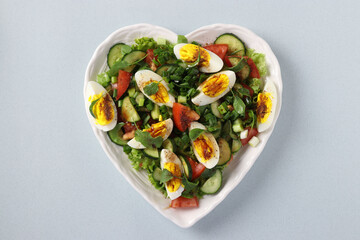 Heart-shaped plate with healthy salad with tomatoes, cucumbers, eggs, arugula and green onion on light blue background, View from above