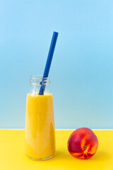 Bright yellow turmeric smoothie in a glass jar with a straw for cocktail, ripe nectarine on a yellow, sky blue background.Healthy nutrition, vegetarianism, detox.A breakfast drink.Refusal of plastic.