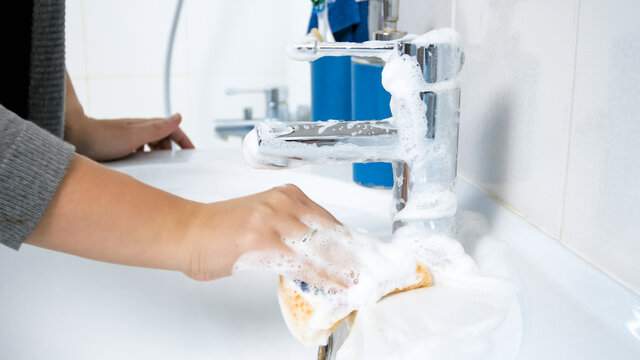 Closeup image of young woman washing bathroom sink with detergent foam and sponge