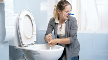 Young housewife cleaning toilet with disgust and closing her nose with clothespin