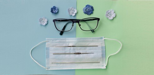 Face mask, eyeglasses, crochet flowers and crochet hooks on blue and green background. Concept of social distancing during covid pandemy.