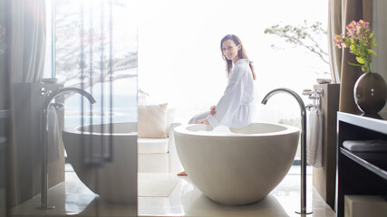 Portrait smiling woman wearing white bathrobe, sitting on edge of bathtub in bathroom - obrazy, fototapety, plakaty