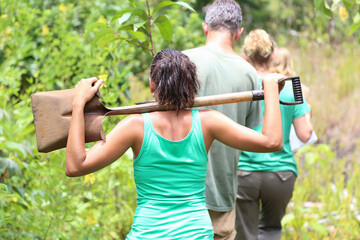 Rear view of woman wearing green tank top carrying shovel on shoulders through garden