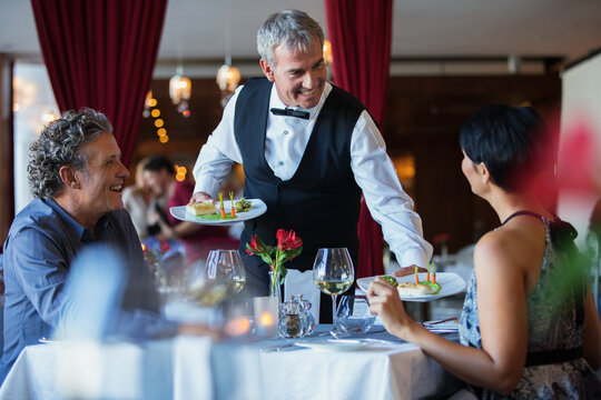 Smiling Waiter Serving Fancy Dishes To Mature Couple Sitting At Table In Restaurant