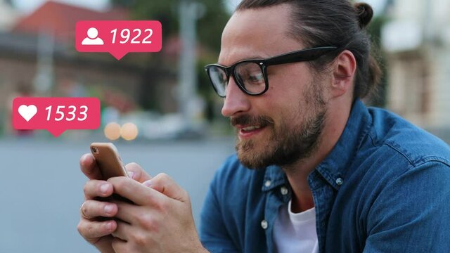 Young Caucasian man in glasses sitting outside in city at street and posting post in social media on smartphone. Guy blogger gathering likes and followers online. Male vlogger liking on mobile phone.