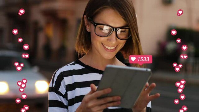 Beautiful Caucasian young woman in glasses standing outdoor at street in evening and liking posts online on tablet device. Female blogger posting in social media and getting reactions icons.