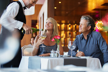 Mature couple sitting at restaurant table, waiter pouring white wine into woman's glass