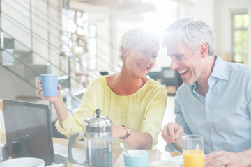 Older couple laughing together at breakfast table with laptop